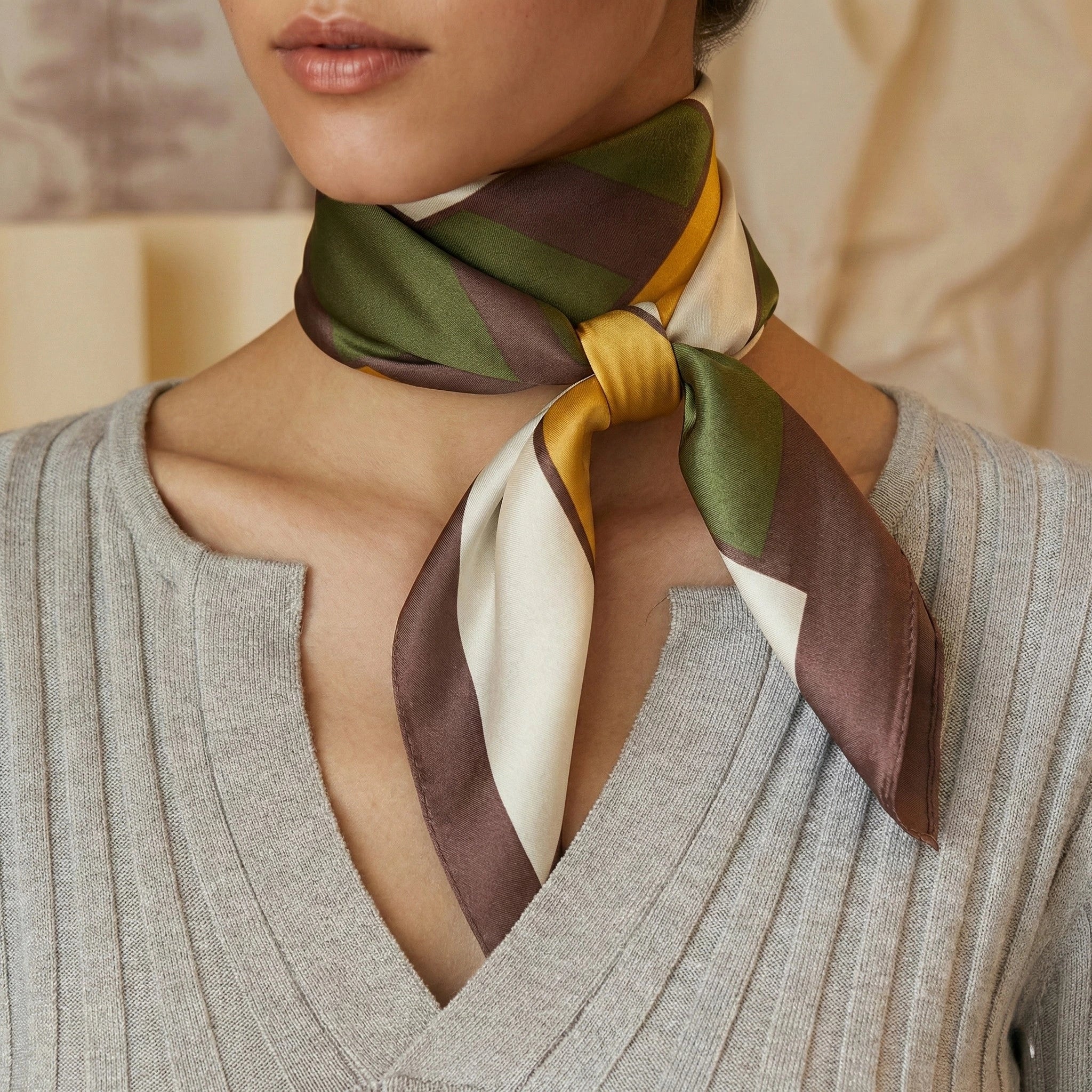 Close-up of a woman wearing an earth-tone geometric silk scarf tied around her neck, styled with a ribbed v-neck sweater Image